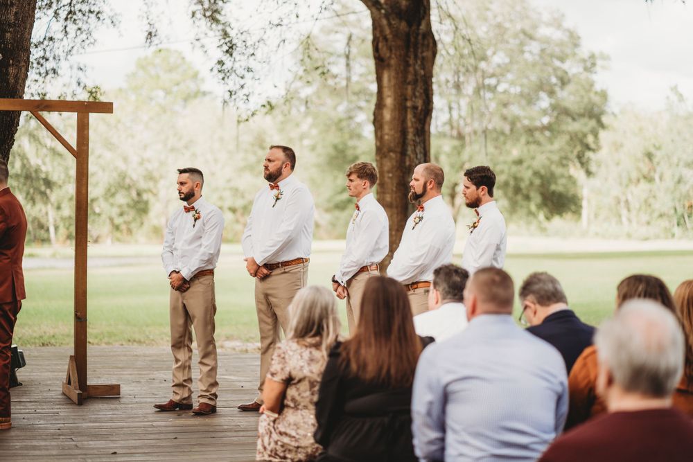 Groomsmen stand at rustic wooden wedding arch in outdoor garden ceremony.