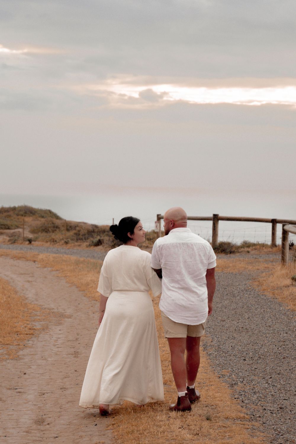 Couple walks hand in hand on a coastal path during sunset.