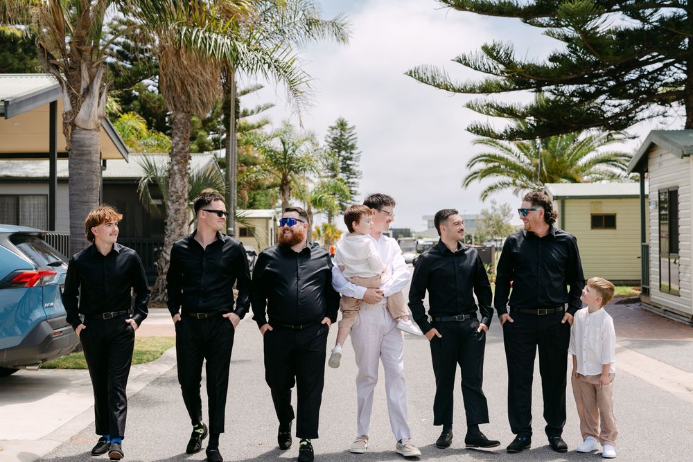 A wedding party in formal attire poses together outdoors on a palm tree-lined path.