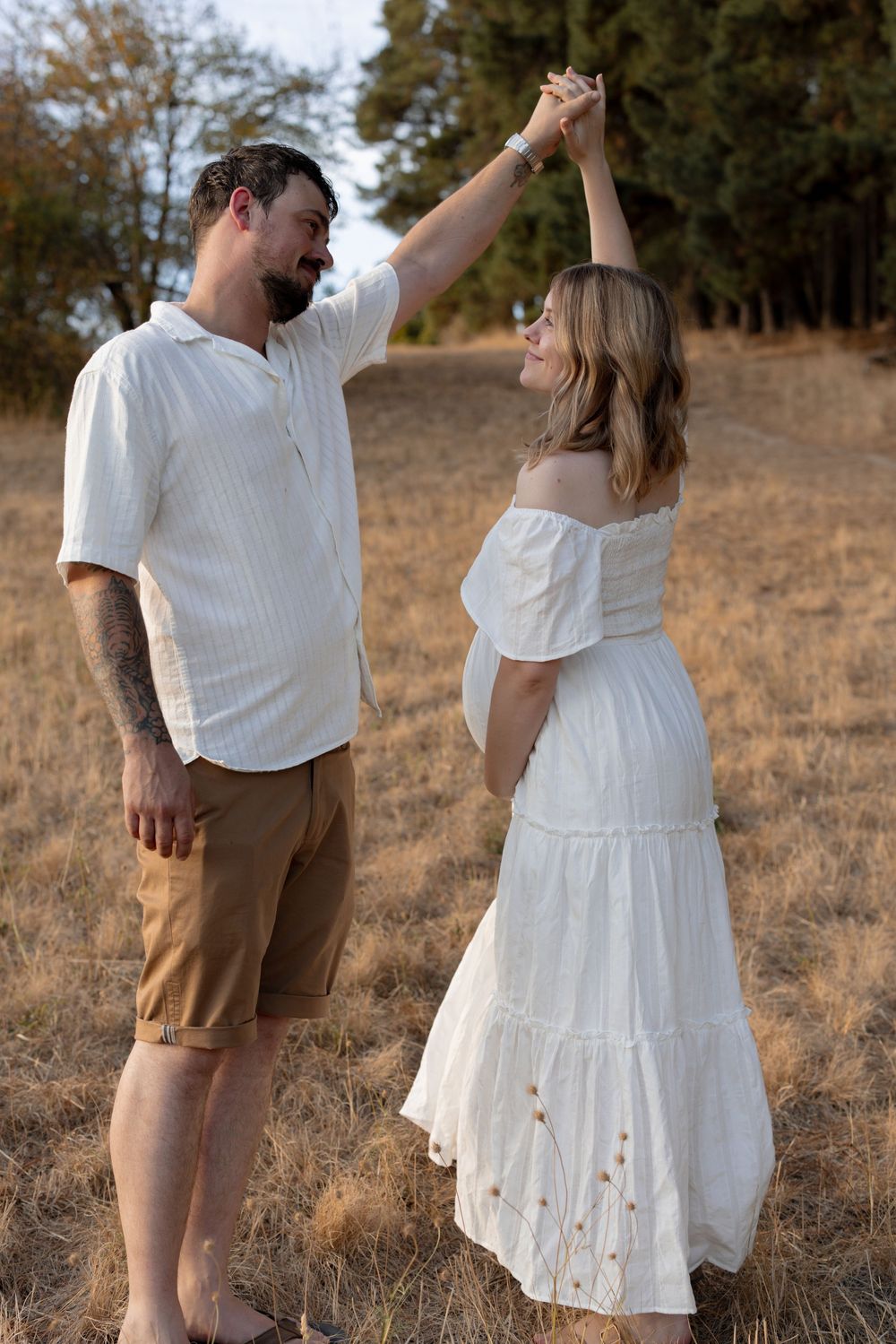 Couple in white and brown outfits dancing together in a grassy field during golden hour.