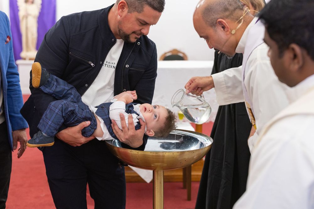 A child being held during a religious baptism ceremony while clergy performs the traditional water blessing ritual.