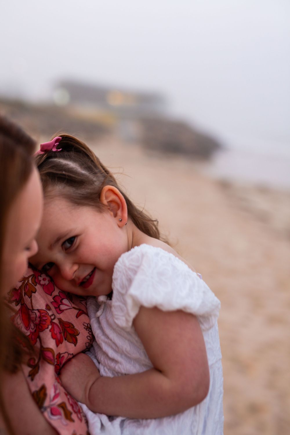 Young child in white dress smiling and cuddling on beach at sunset.