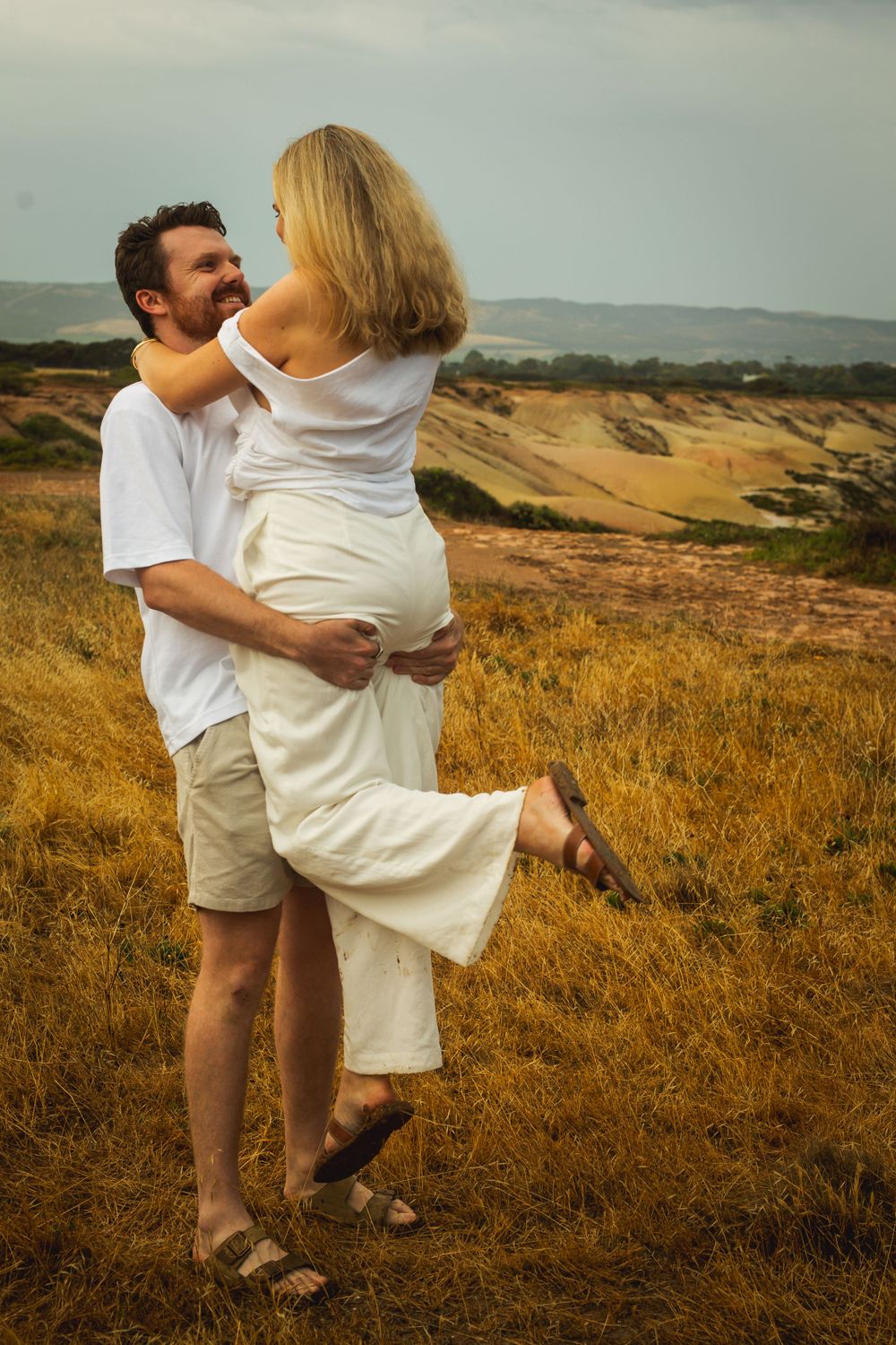 Romantic couple embracing in golden field during sunset with rolling hills in background.