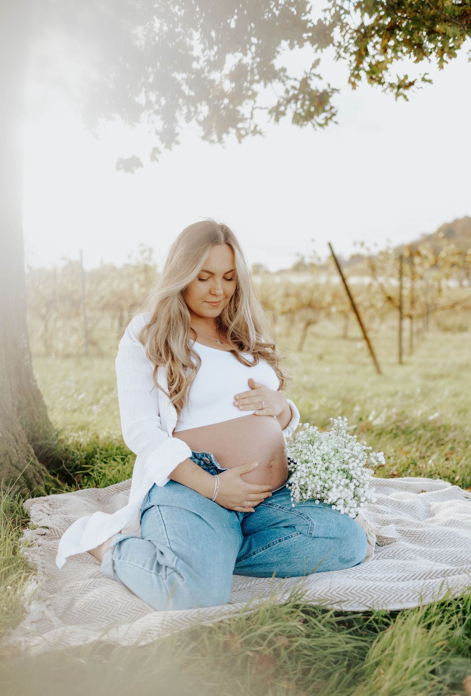 Expectant figure in white top and jeans sitting on blanket outdoors holding small white flowers.