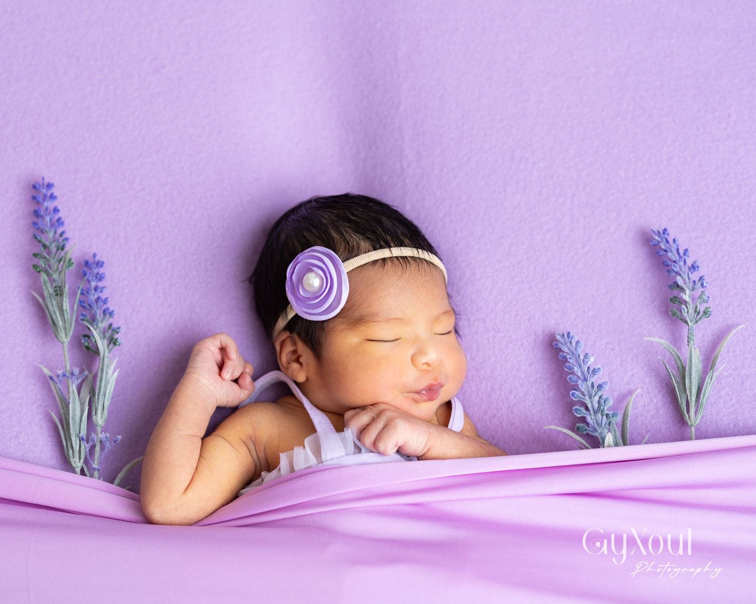 Sleeping newborn baby posed on purple fabric with lavender sprigs and a floral headband.