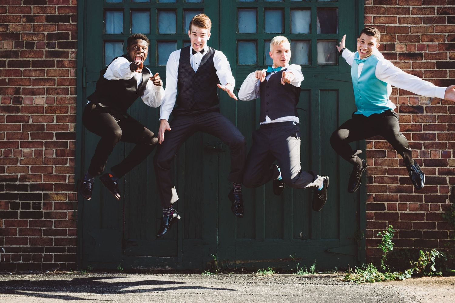 Four groomsmen leap joyfully against a brick wall backdrop.