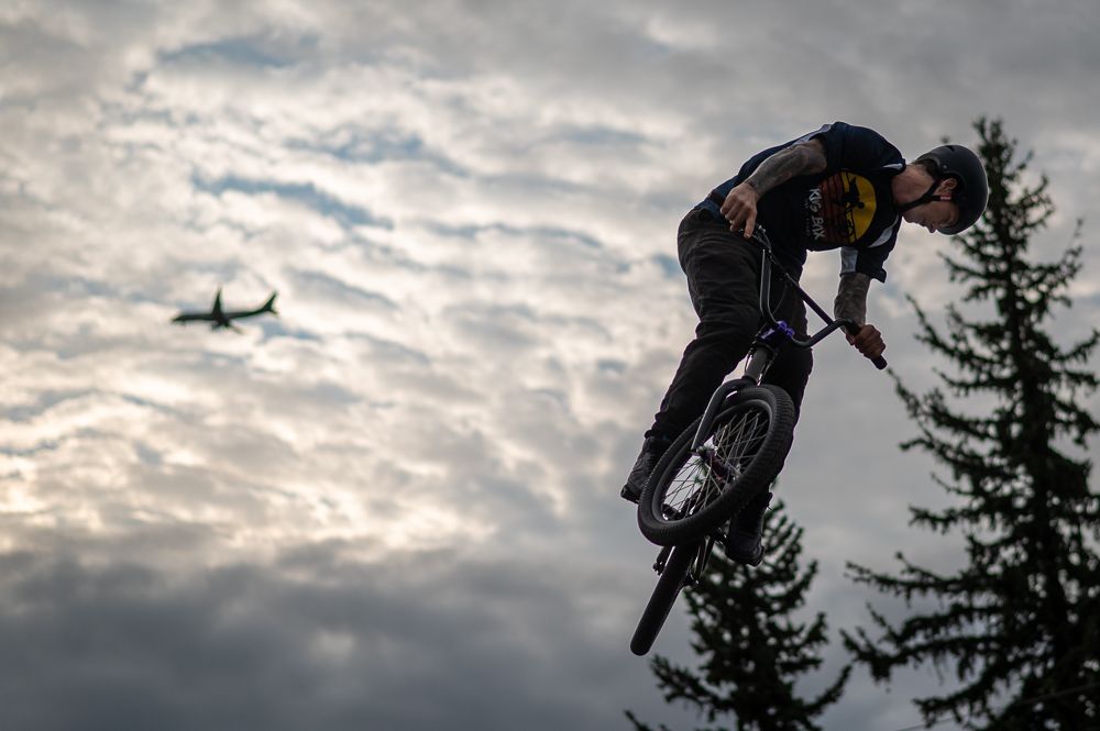 BMX rider performs aerial jump against dramatic cloudy sky with airplane in background.