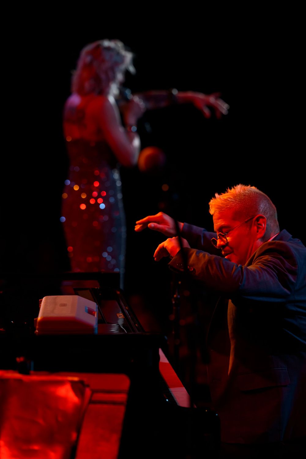 Musician plays piano on stage under red light while a female singer performs in a sequined dress in the background.