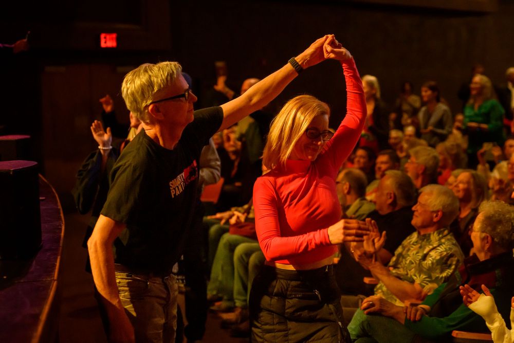 Couple dancing together at a live event, woman in red top being twirled by man in black shirt, audience seated behind them.