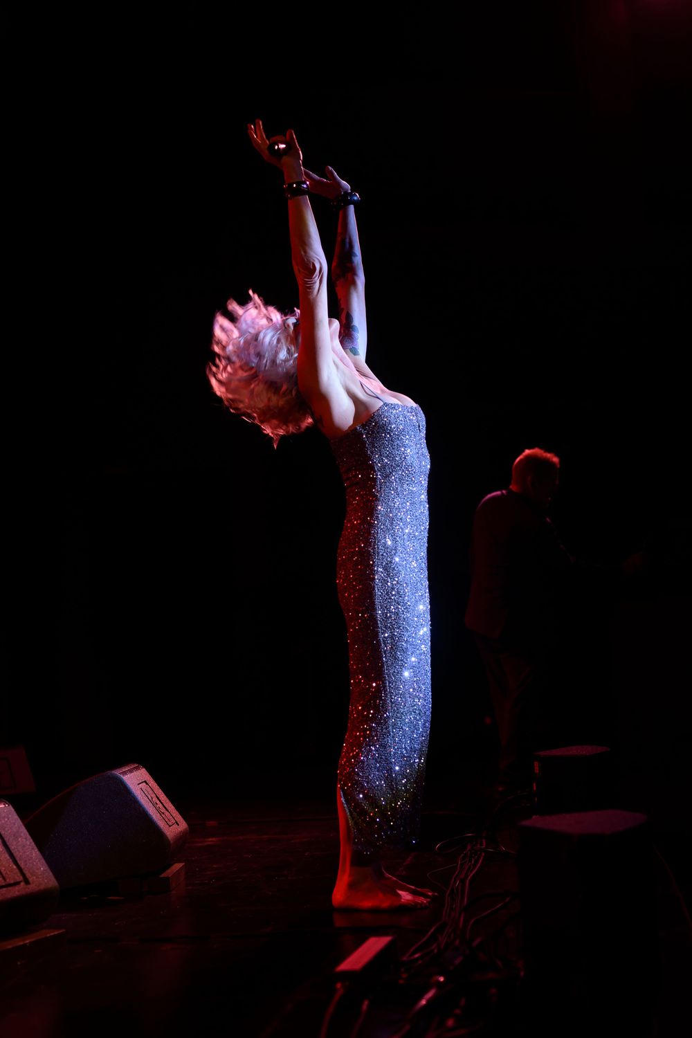 A performer in a sparkling silver gown stretches arms upward on a dark stage under dramatic lighting.
