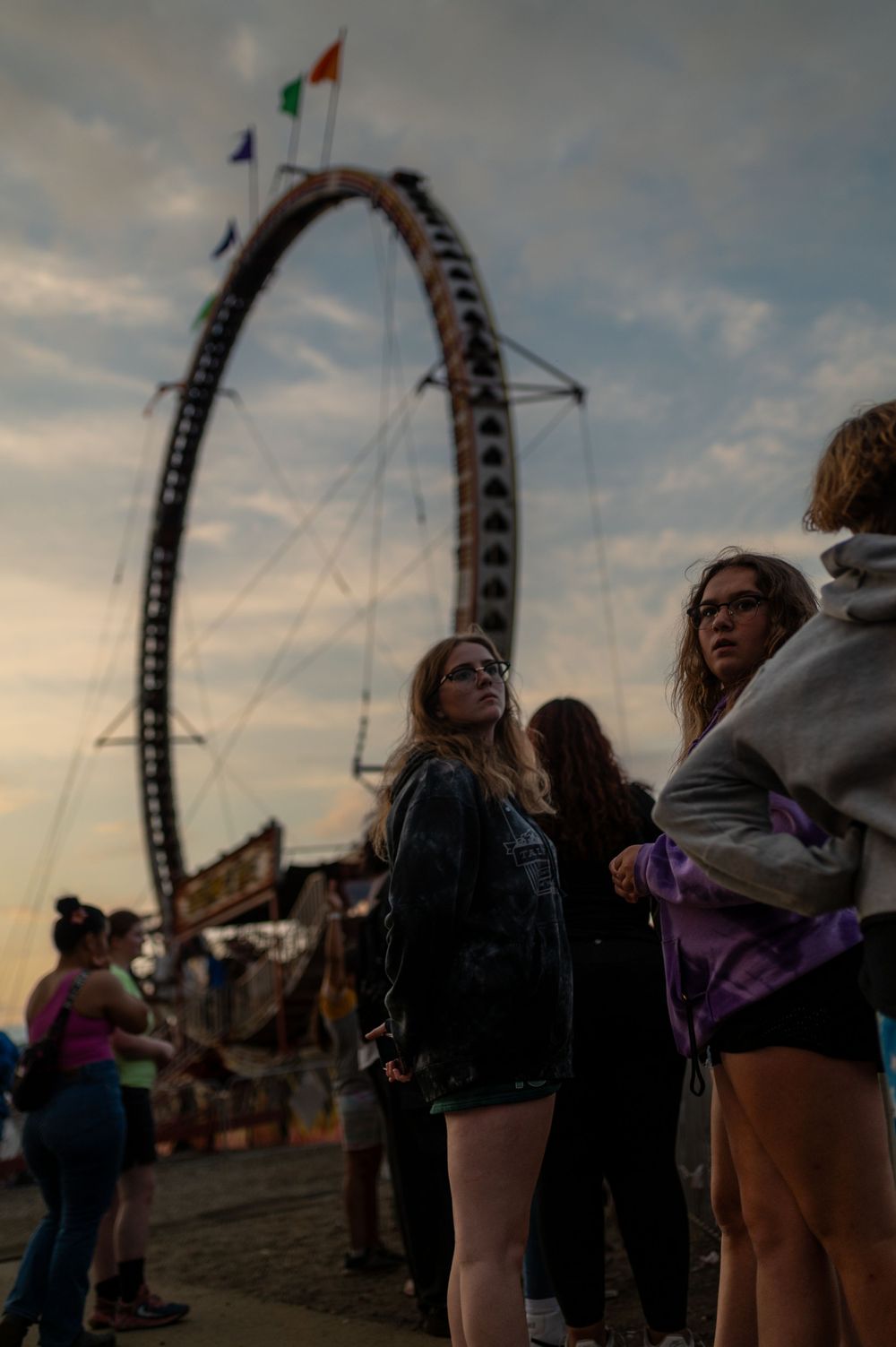 Silhouettes of fairgoers against a dramatic sunset with a roller coaster loop in the background.