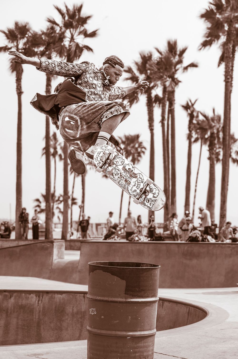 Skateboarder performing a high kickflip trick at Venice Beach skate park with palm trees in sepia tone.