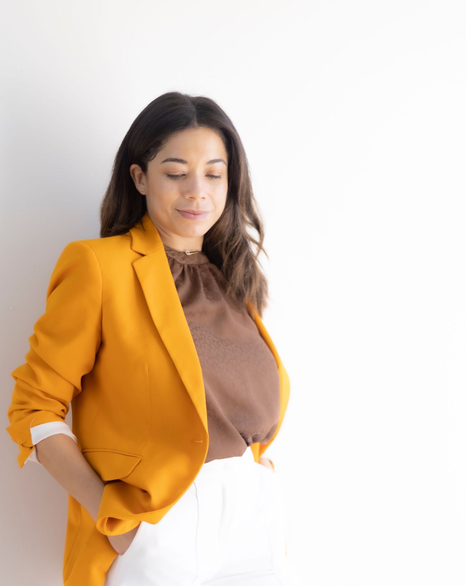 Fashion portrait showcasing yellow blazer and brown top combination against white wall.