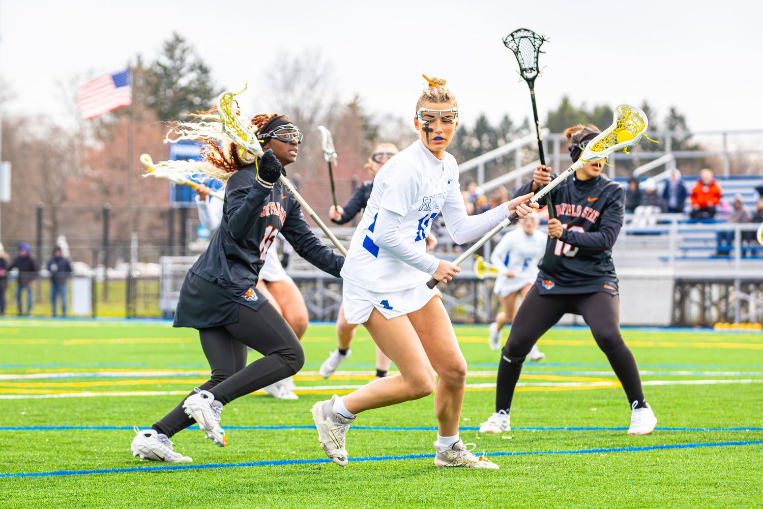 Lacrosse players engage in a dynamic play sequence on an artificial turf field during a competitive match.