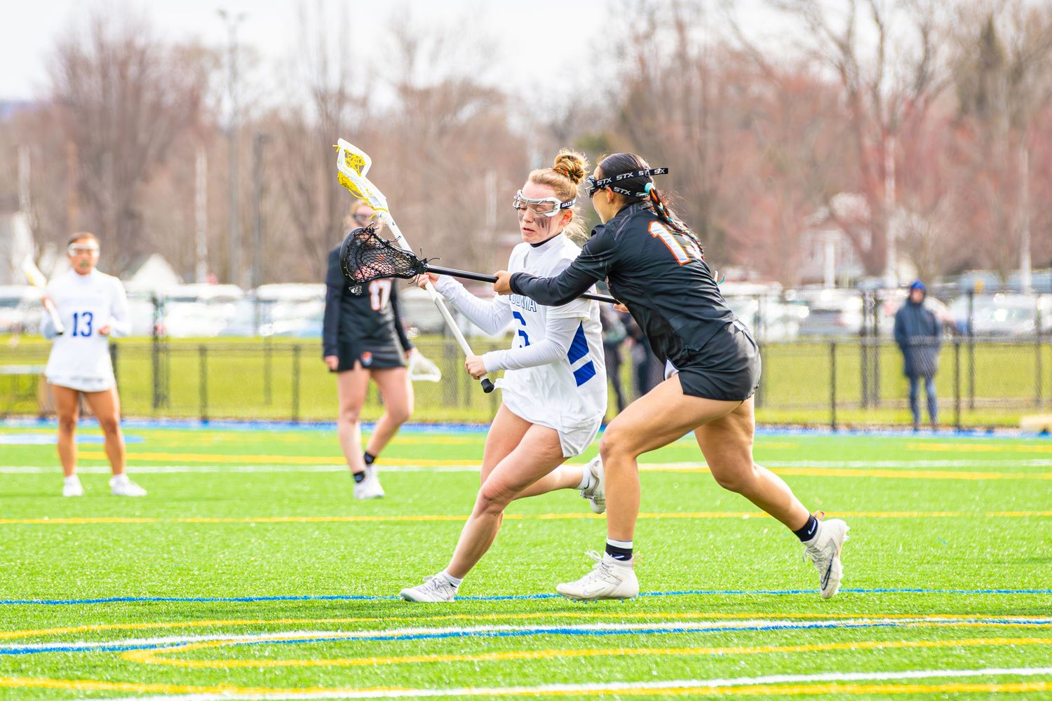 Athletes compete intensely during a lacrosse match on a grass field with spectators watching from the sidelines.