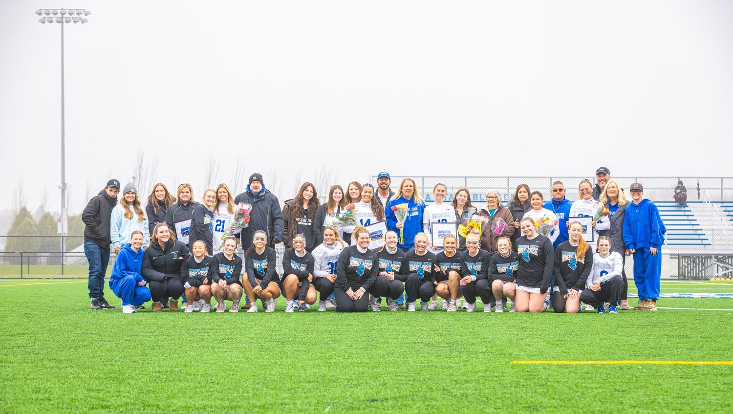 Large group of athletes and spectators gather on the sidelines of a sports field.