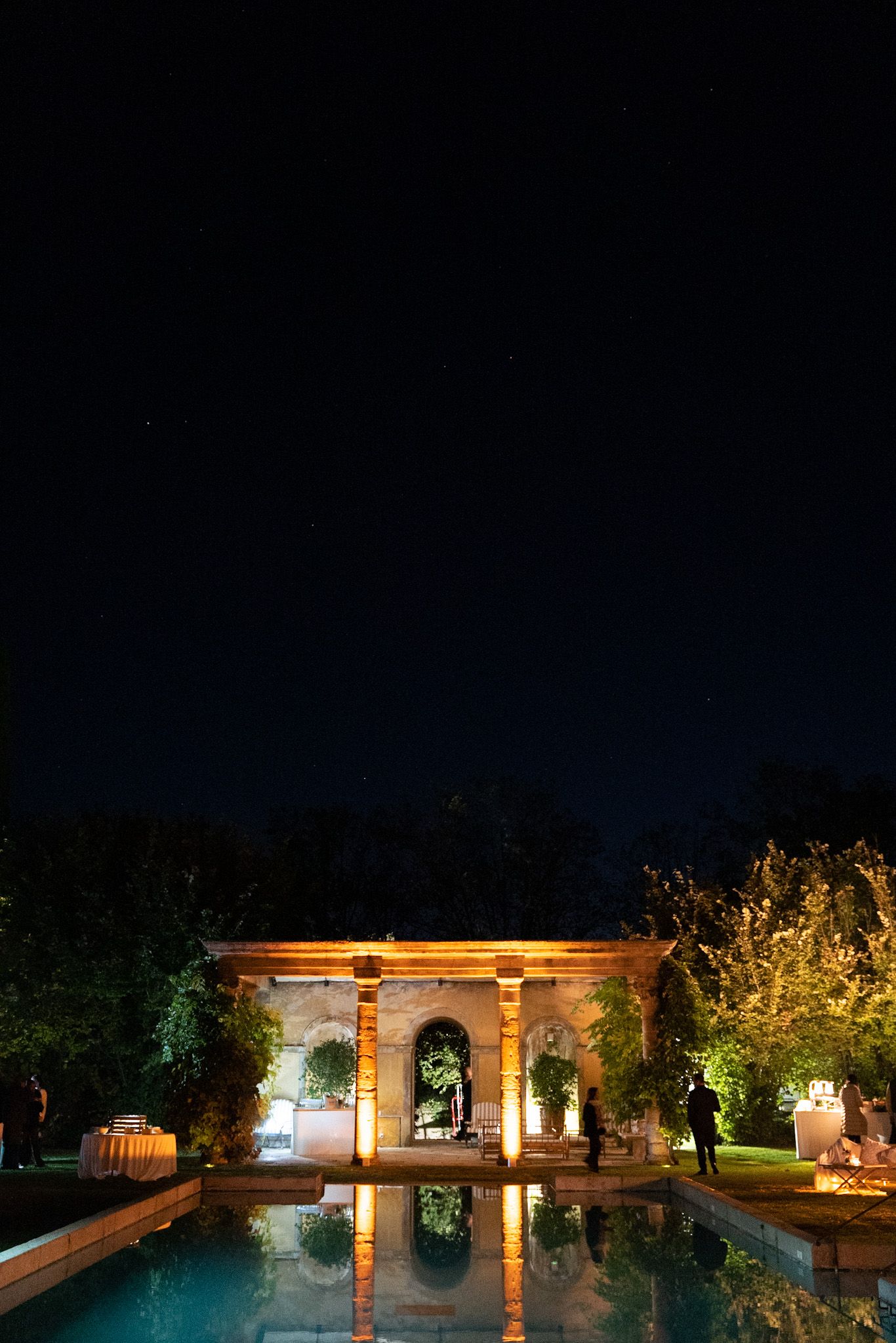 Illuminated stone pavilion and reflecting pool at night with architectural columns and landscaped gardens.