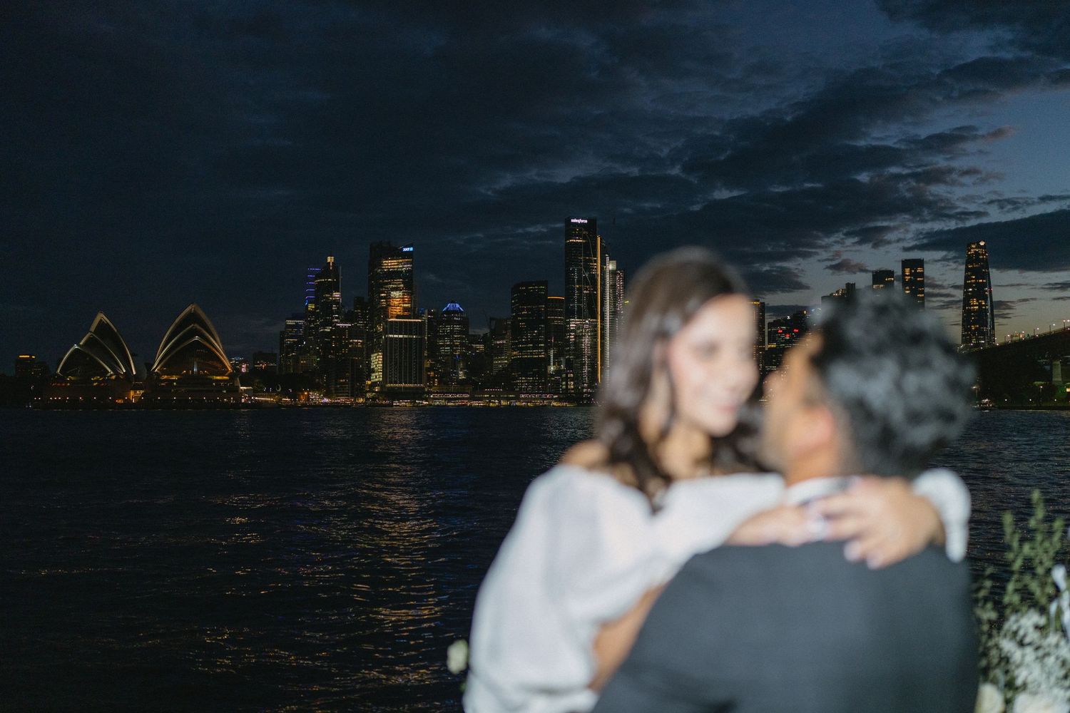 Sydney Opera House and harbor skyline illuminated at dusk with silhouetted couple in foreground.