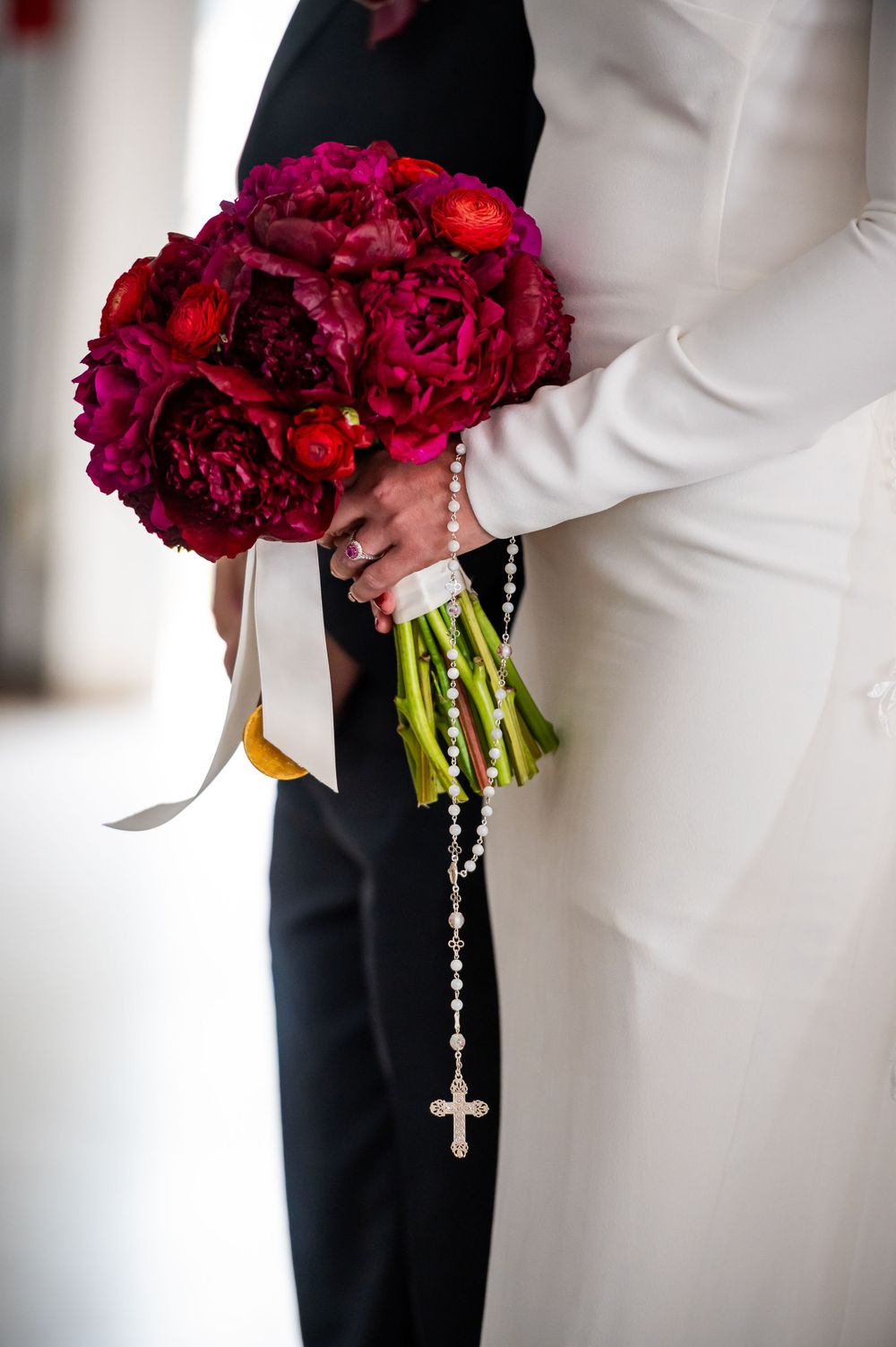 Deep red peony bouquet with rosary beads held against white wedding dress.