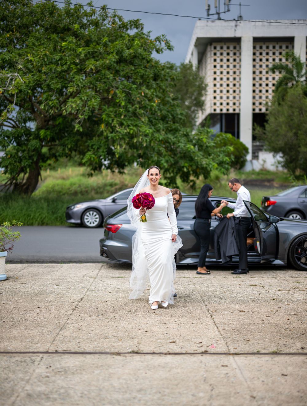 Bride in elegant white dress walks outside church holding red bouquet.