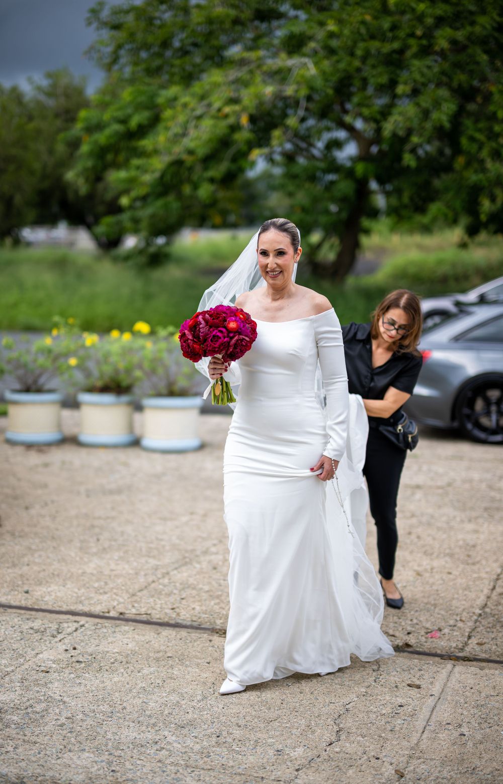 Bride in off-shoulder wedding gown walks outdoors while holding red bouquet.