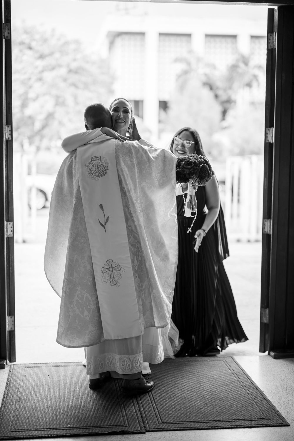 Black and white photo of priest greeting wedding party at church entrance.