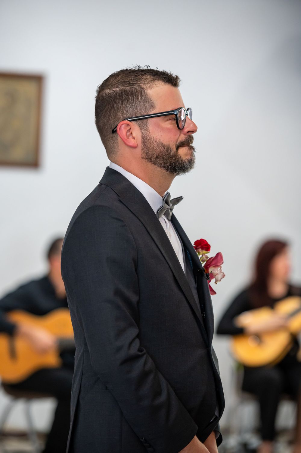 Groom wearing glasses and black suit stands at altar with guitarist in background during wedding ceremony.