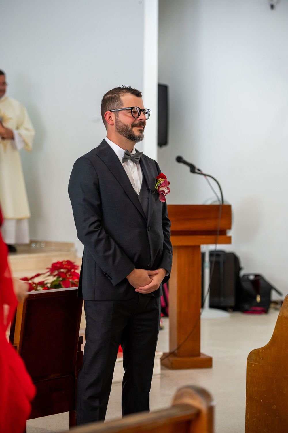 Groom in dark suit and bow tie stands at church podium during wedding ceremony.