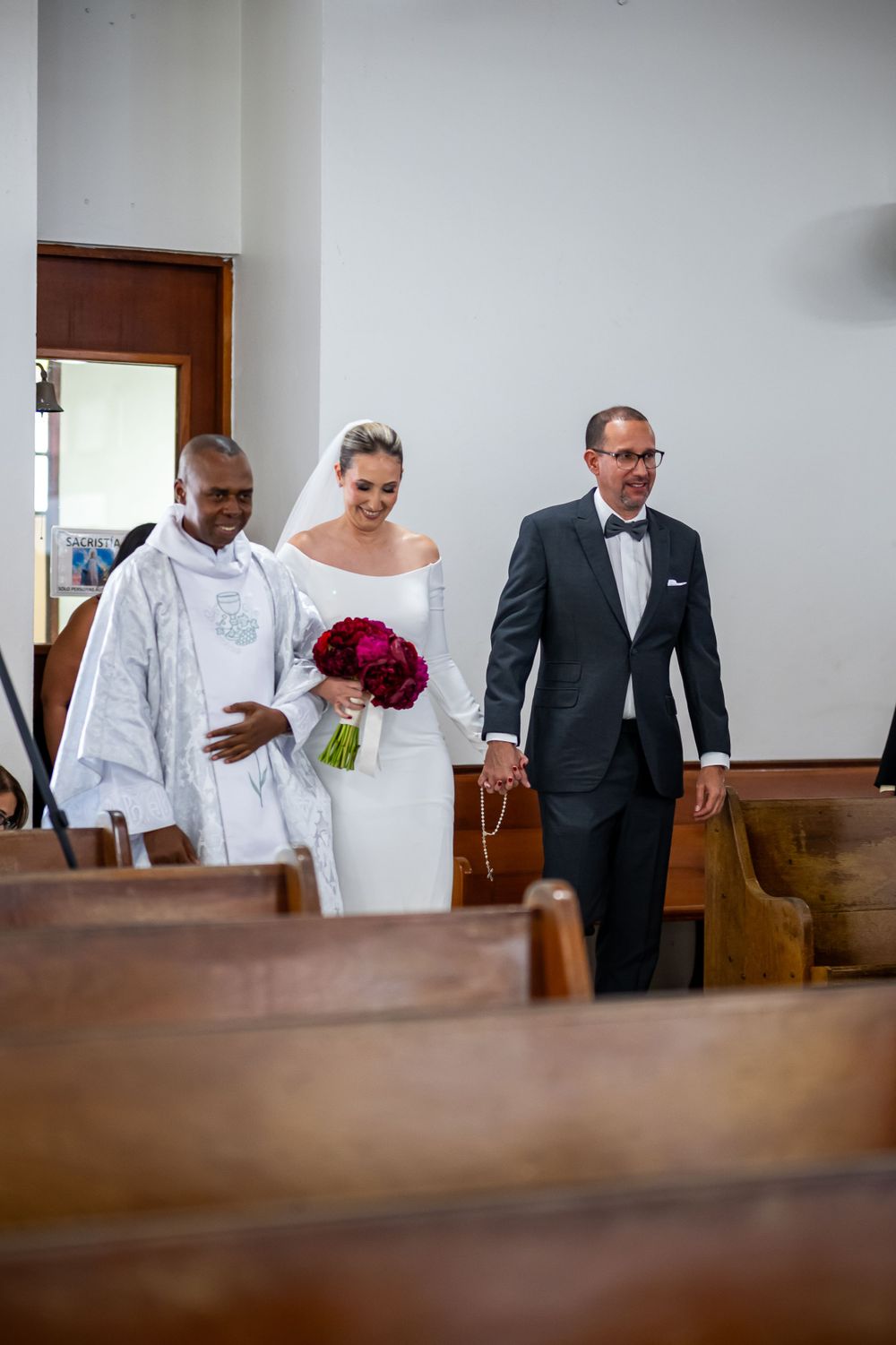Wedding party processes down church aisle with bride holding deep red bouquet.