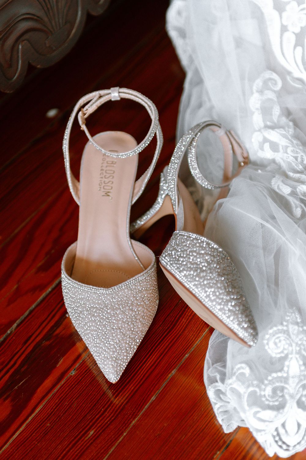 Sparkly silver bridal shoes next to white lace wedding dress on red wooden floor.