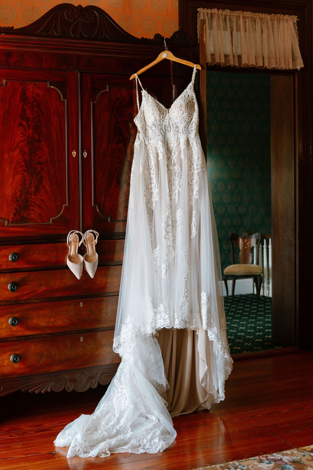 A white lace wedding dress hangs from a wooden wardrobe against green wallpaper.