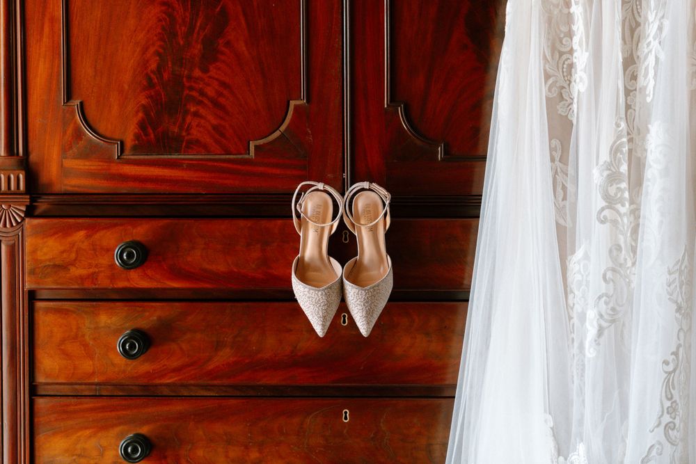 Elegant white pointed-toe bridal shoes with sparkles hanging against a wooden dresser drawer.