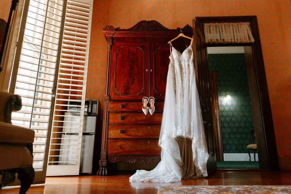 A white wedding dress hangs beside a full-length mirror in a traditional room with wooden furniture.