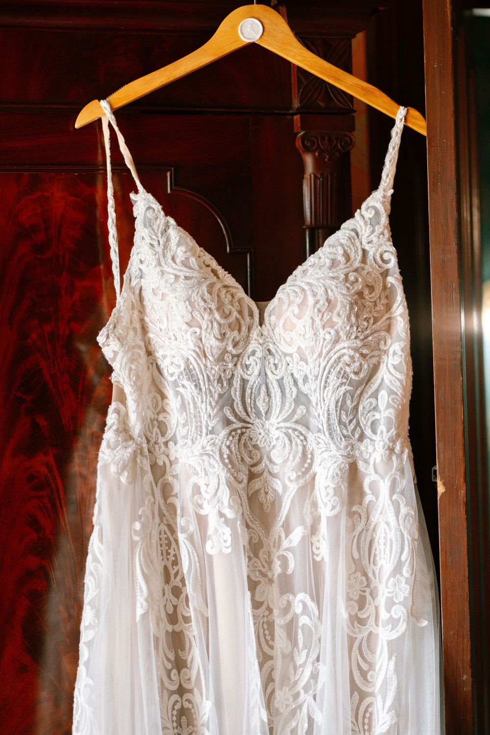 Close-up of an intricate lace wedding dress bodice with spaghetti straps hanging on a wooden hanger.