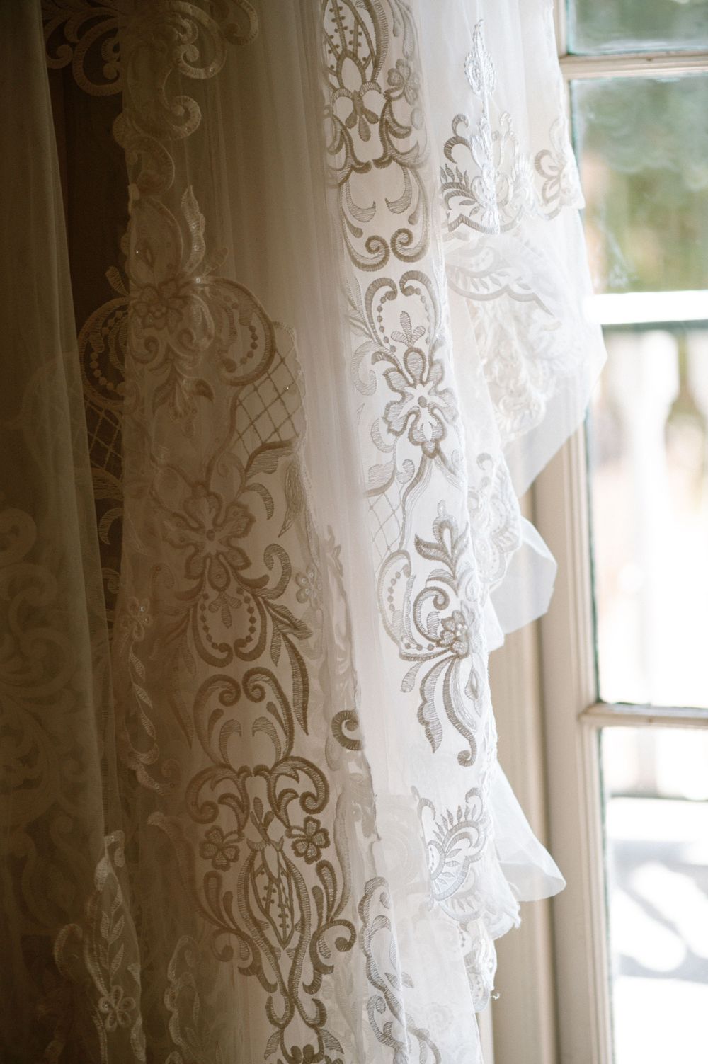 Close-up of delicate lace embroidery details on white wedding dress fabric near a window.