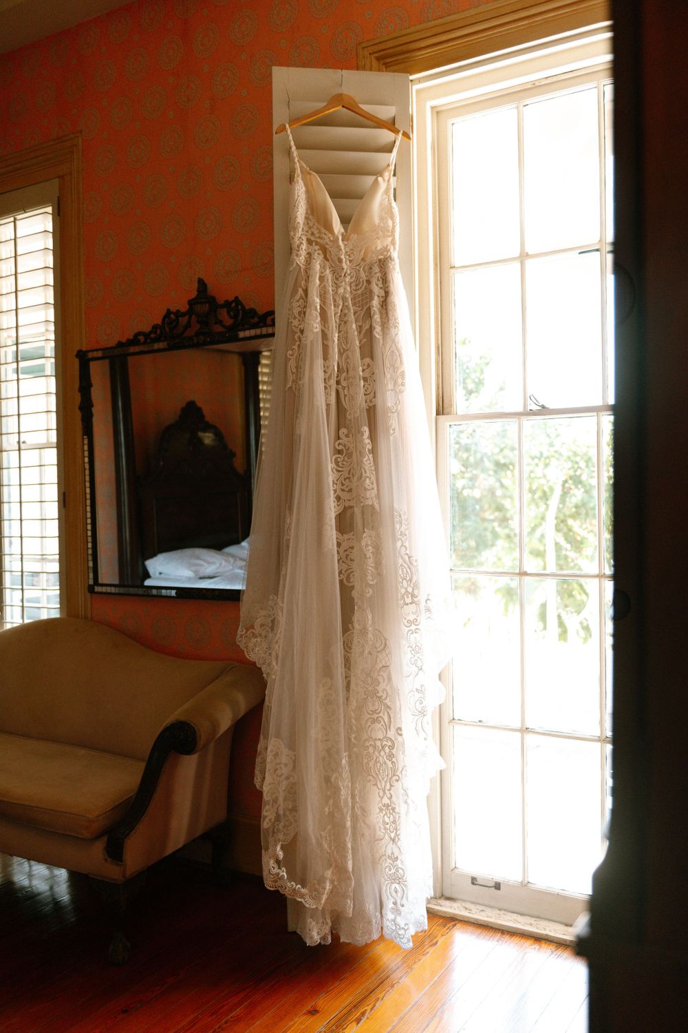Sunlit wedding gown hanging by a tall window in an elegant room with antique furniture.