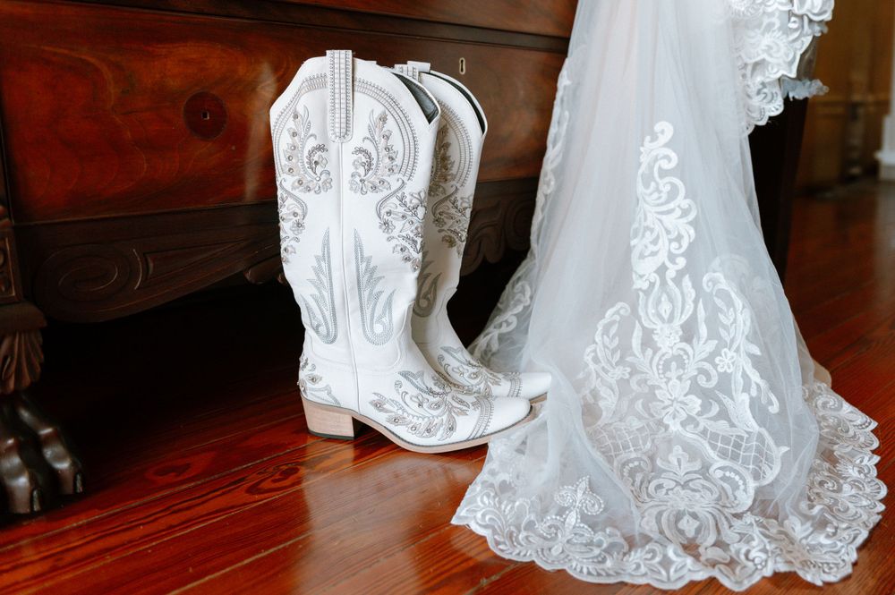 White cowboy boots next to the hem of a lace wedding dress on hardwood flooring.