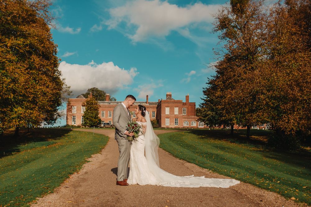 Newlyweds pose on path leading to brick mansion with autumn trees framing the scene.