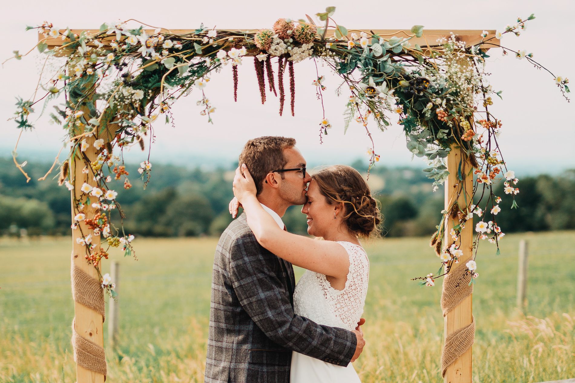 Wedding couple embrace under rustic wooden arch decorated with flowers and greenery in countryside setting.