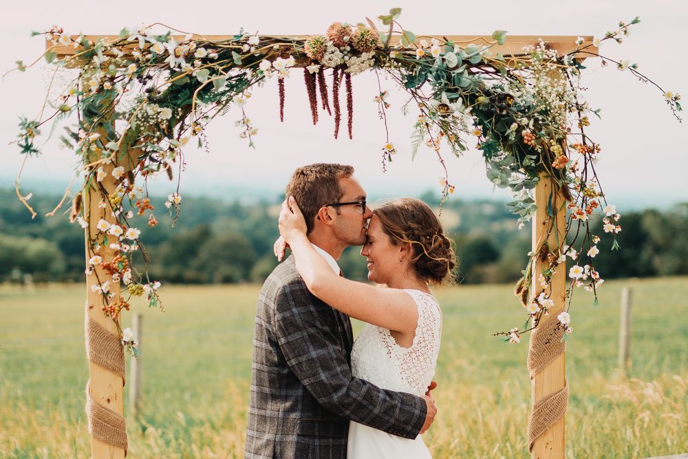 Wedding couple embrace under rustic wooden arch decorated with flowers and greenery in countryside setting.