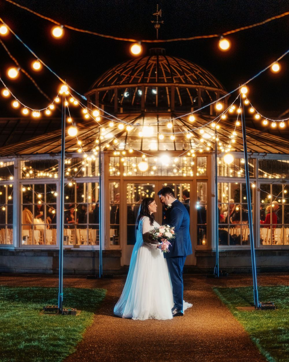 A couple stands under a beautifully lit gazebo with string lights creating a romantic evening atmosphere at Chiswick House Gardens.