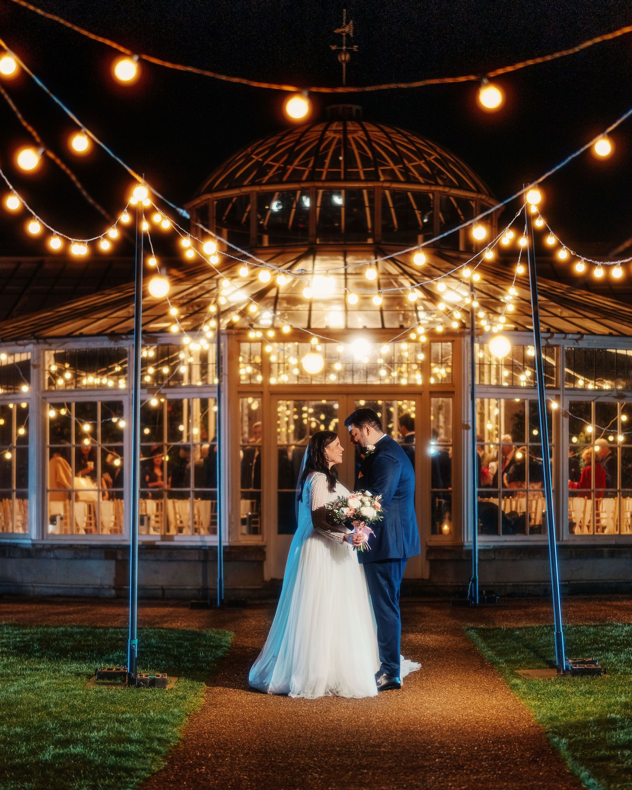 A couple stands under a beautifully lit gazebo with string lights creating a romantic evening atmosphere at Chiswick House Gardens.