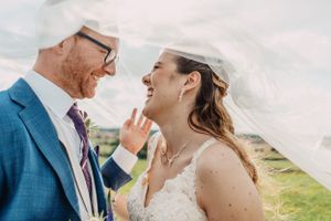 Candid moment of laughter shared under flowing white veil against countryside backdrop.