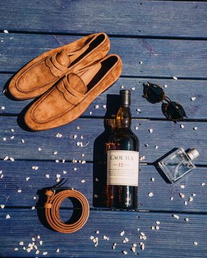Brown leather loafers, sunglasses, and whiskey bottle arranged on blue wooden deck with scattered petals in Crete.