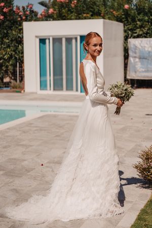 An elegant bride in a long-sleeved white wedding gown holds a bouquet by a modern poolside in Crete.