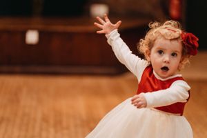 Child in white tulle dress with red sash dances joyfully on wooden floor.