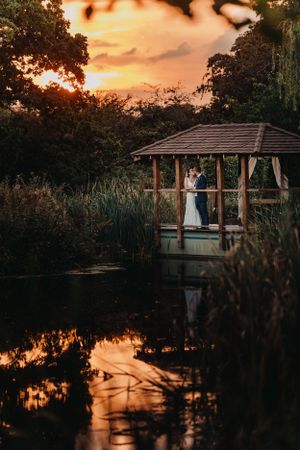 Romantic sunset wedding photo in wooden gazebo overlooking reflective pond.