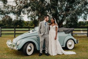 Newlyweds pose beside classic light blue vintage Volkswagen Beetle on grassy field at Ham Polo Club.