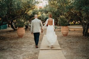 A wedding couple walks hand in hand down a path lined with olive trees and terracotta pots in Crete.
