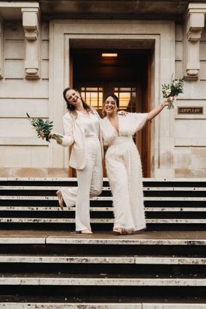 Two people in matching white outfits pose joyfully on stone steps of the historic Hackney Town Hall.